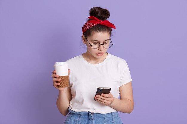 Morena atraente, jovem caucasiana, segura um celular moderno e café para viagem, envia mensagens de texto no chat online, usa uma camiseta casual branca e uma faixa vermelha