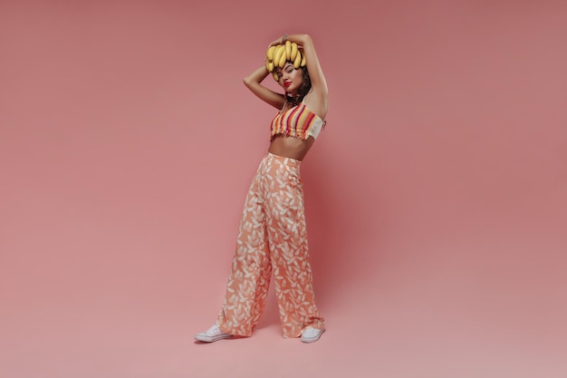 Morena elegante com batom brilhante no top colorido e calça larga rosa olhando para a câmera e segurando bananas
