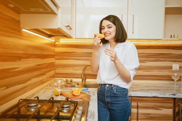 Morena jovem e bonita em pé na cozinha com laranja