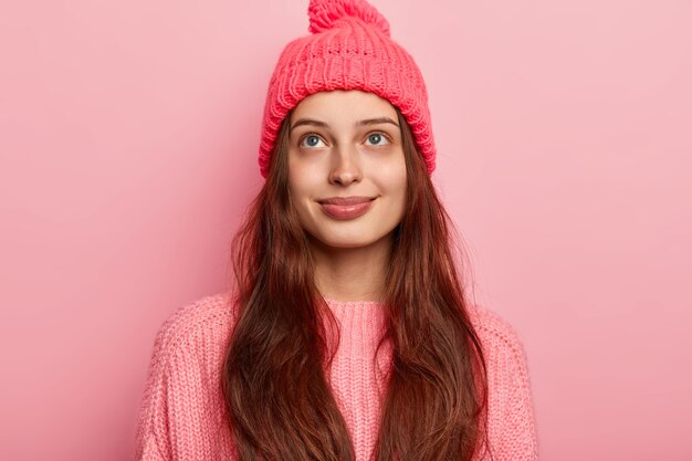 Morena sonhadora satisfeita, menina milenar, olha para cima com pensamentos agradáveis, usa um suéter e chapéu de malha vívidos, modelos sobre fundo rosa, tem um sorriso charmoso e um olhar terno