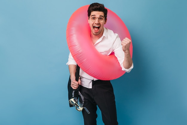 Moreno homem de calça preta e camisa branca grita com emoção e alegria. Cara de óculos segura bolsa preta, máscara para nadar e coloca um círculo inflável.