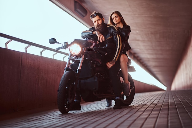 Motociclista barbudo brutal na jaqueta de couro preta e menina morena sensual sentados juntos em uma motocicleta retrô feita sob medida em uma passarela debaixo de uma ponte.