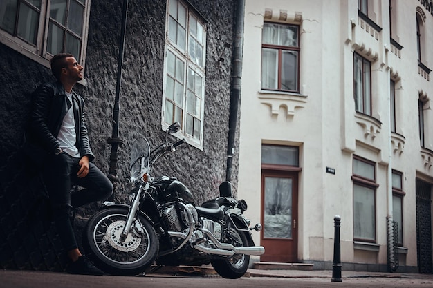 Motociclista elegante vestido com uma jaqueta de couro preta e jeans encostado em uma parede perto de sua motocicleta retrô em uma velha rua da europa.