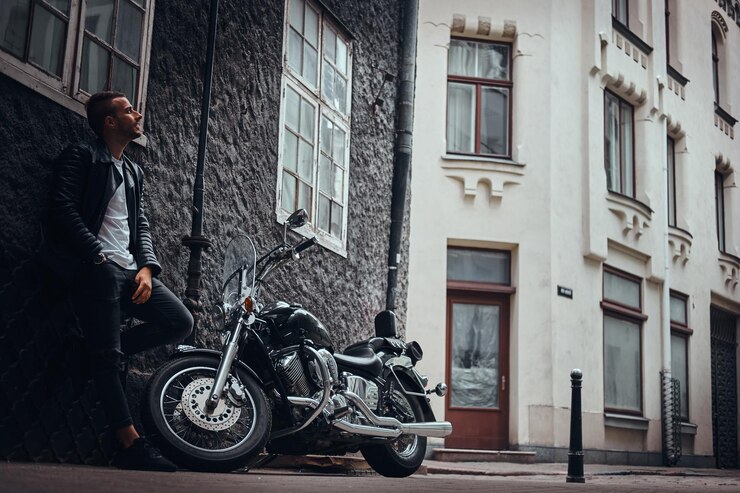 Foto grátis motociclista elegante vestido com uma jaqueta de couro preta e jeans encostado em uma parede perto de sua motocicleta retrô em uma velha rua da europa.