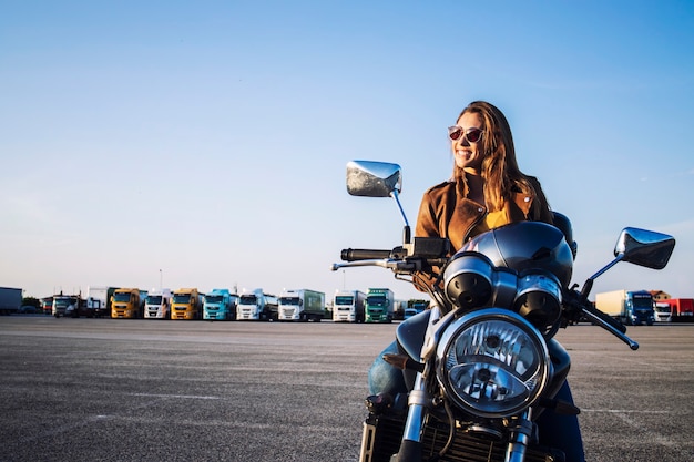 Motociclista feminina em uma jaqueta de couro, sentada em uma moto retrô e sorrindo Foto gratuita