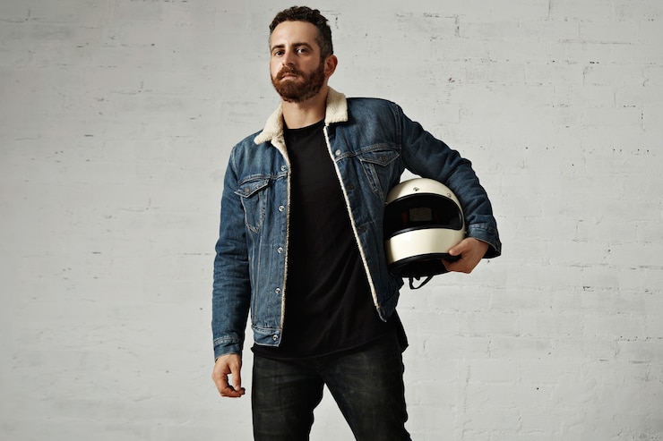 Foto grátis motociclista usa jaqueta jeans shearling e camisa branca henley preta, segura um capacete bege vintage para motociclista, isolado no centro da parede de tijolos brancos