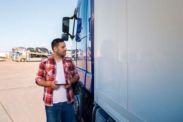 Motorista de caminhão com roupas casuais parado ao lado de seu caminhão com um tablet e olhando para o caminhão