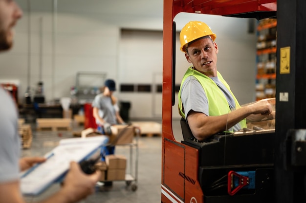 Motorista de empilhadeira sorridente se comunicando com o despachante sobre detalhes da carga no armazém industrial