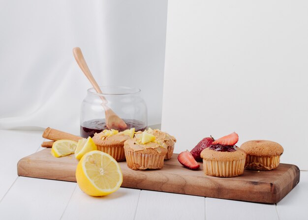 Muffins de vista lateral com morangos e limão em um quadro negro com um pote de geléia e um caderno branco