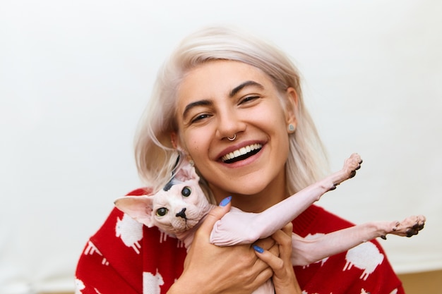 Muito adolescente com cabelo rosado e piercing no nariz, abraçando o gato Sphynx e sorrindo. Mulher europeia elegante e bonita acariciando seu gatinho, posando isolada, com expressão facial alegre