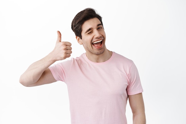 Muito bom mano. homem sorridente e piscando mostra polegares em aprovação, gosta e apóia sua escolha, elogia o ótimo trabalho, excelente trabalho, ficando satisfeito contra um fundo branco