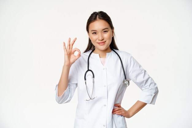 Muito bom sorridente e confiante médica asiática mostrando tudo bem, ok, sinal de aprovação, confirmar smth dizendo ...