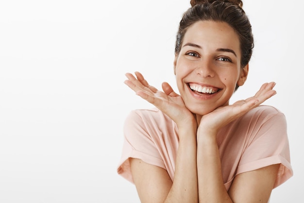 Muito confiante e gentil jovem elegante com gesto de coque cuidando da pele sentindo-se feliz se livrando das imperfeições inclinando a cabeça nas palmas das mãos e sorrindo amplamente como regozijando-se com o bom resultado