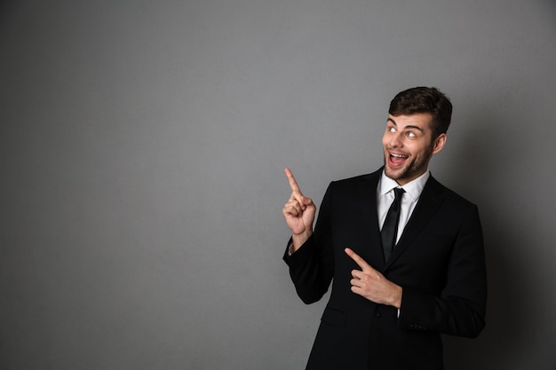 Muito feliz homem morena de terno clássico, apontando com dois dedos, olhando de lado