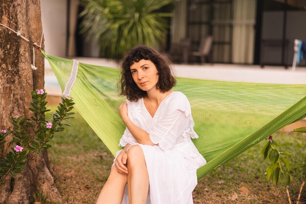 Muito jovem morena caucasiana vestindo vestido branco olha para a câmera sentada na rede no jardim durante o dia