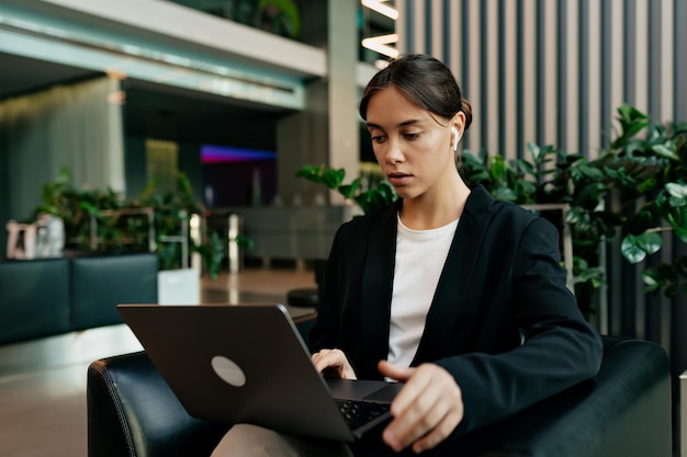 Muito jovem mulher caucasiana está estudando enquanto está sentado no escritório moderno Mulher atraente em camiseta branca e jaqueta escura passa tempo fazendo atividades úteis Conceito de educação