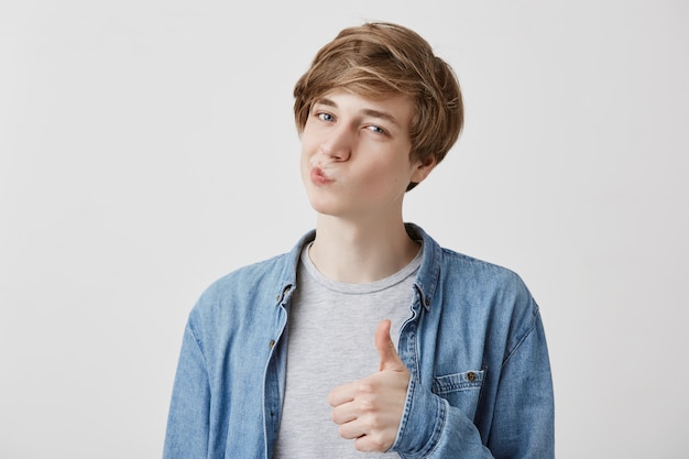 É muito melhor assim. Entusiasmado alegre jovem homem caucasiano em camisa jeans com cabelo loiro, mostrando os polegares para cima gesto para a câmera, fazendo beicinho, expressa sua aprovação da idéia ou projeto