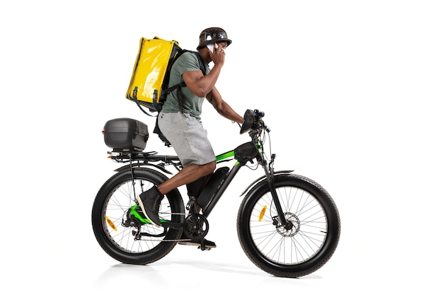 Muitos pedidos. Serviço de entrega sem contato durante a quarentena. O homem entrega comida durante o isolamento, usando capacete e máscara facial. Levando comida na bicicleta isolada na parede branca. Segurança. Se apressando.