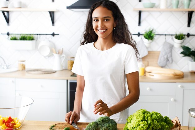 Mulata bonita está sorrindo e segurando uma faca na cozinha moderna, vestida com camiseta branca, perto da mesa com legumes frescos