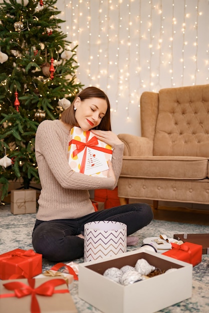 Mulher abraçando uma caixa de presente enquanto está sentada entre muitos presentes na sala de estar decorada de natal