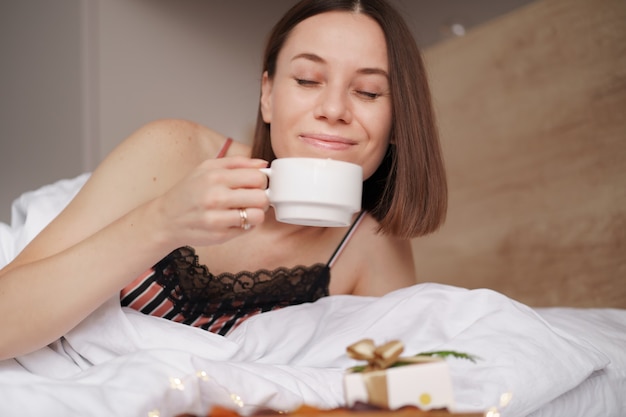 Mulher acordada na cama com um presente e café com marshmallows em pé perto dela.