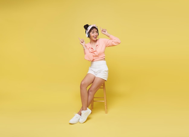 Mulher adolescente asiática feliz sorrindo ouvindo música em fones de ouvido e cadeira sentada em fundo amarelo