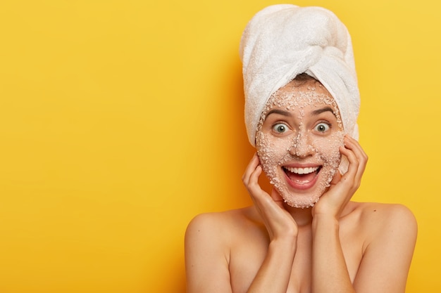 Mulher adorável alegre com expressão facial alegre, aplica máscara natural de sal marinho, tem pele saudável e bem cuidada, usa toalha na cabeça, gosta de tratamentos higiênicos