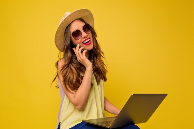 Mulher adorável e eficaz usando chapéu e vestido de verão, andando ao telefone e trabalhando com o laptop