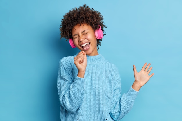 Mulher adorável e otimista tem cabelo encaracolado ouve música em fones de ouvido sem fio, canta junto levanta as mãos usa um suéter casual empolgado com a música favorita vestida casualmente