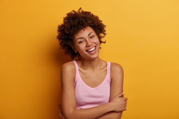 Mulher adorável feliz e despreocupada de pele escura mantém as mãos cruzadas sobre o corpo, sorri suavemente, tem humor otimista, tem cabelos cacheados naturais, desfruta de momentos agradáveis na vida, isolada na parede amarela