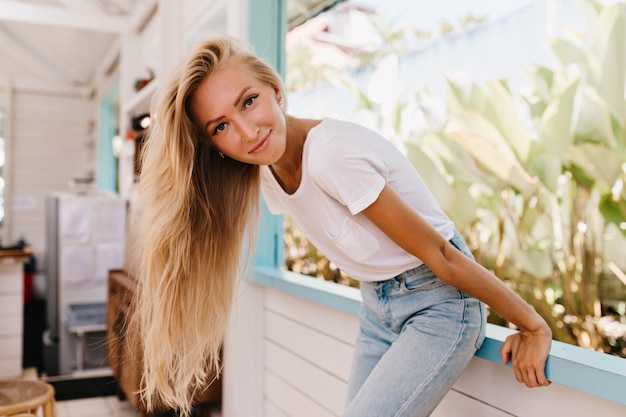 Mulher adorável graciosa com penteado longo, posando perto da janela. adorável garota de cabelos louros sentado no peitoril e rindo.