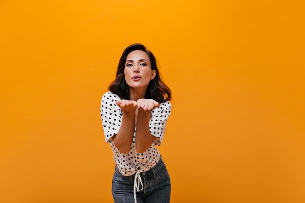 Mulher adorável manda beijo em fundo laranja. Mulher de cabelos escuros em poses de blusa de bolinhas brancas e jeans azul para a câmera.