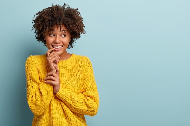 Mulher adorável otimista com cabelos cacheados, mantém a mão perto de um sorriso dentuço, olha para o lado, tem expressão sonhadora, sente-se tímida, usa um macacão amarelo casual, fica de pé contra a parede azul. Copie o espaço certo