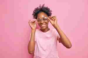 Foto grátis mulher adorável positiva com pele escura de cabelo encaracolado toca a borda dos óculos sorri com dentes olha diretamente para a câmera alegremente vestida com camiseta casual isolada sobre fundo rosa conceito de emoções