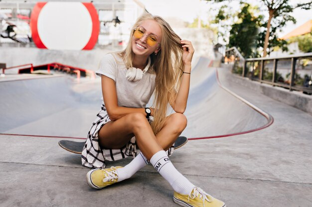 Mulher adorável skatista brincando com seu cabelo loiro. Retrato ao ar livre da linda modelo feminino em sapatos amarelos, sentado no skate.