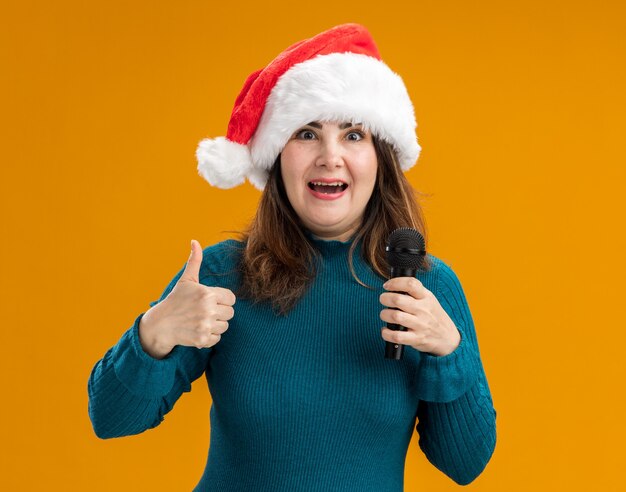 Mulher adulta caucasiana animada com chapéu de Papai Noel com os polegares para cima e segurando o microfone isolado em um fundo laranja com espaço de cópia