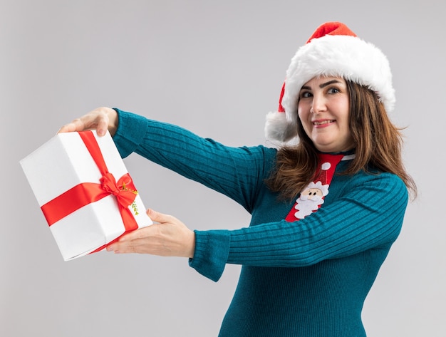 Mulher adulta caucasiana feliz com chapéu de Papai Noel e gravata de Papai Noel segurando uma caixa de presente de Natal, olhando para a câmera, isolada no fundo branco com espaço de cópia