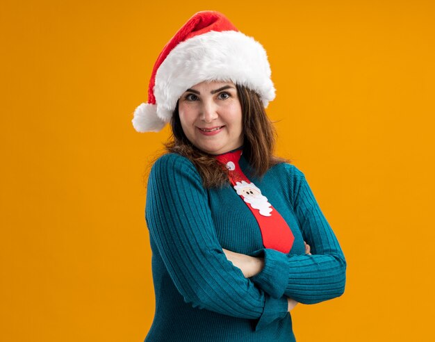 Mulher adulta caucasiana sorridente com chapéu de Papai Noel e gravata de Papai Noel fica com os braços cruzados isolados na parede laranja com espaço de cópia