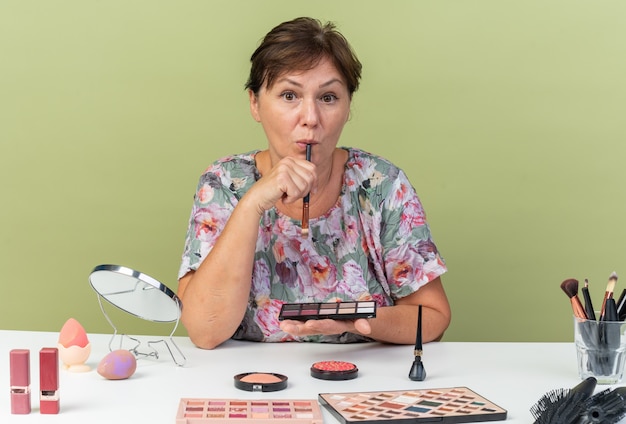 Foto grátis mulher adulta caucasiana surpreendida sentada à mesa com ferramentas de maquiagem segurando uma paleta de sombras e um pincel de maquiagem