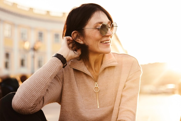 Foto grátis mulher adulta de pele clara sorridente em óculos de sol e roupas casuais sentada na cidade e relaxando o conceito de emoções e estados de espírito