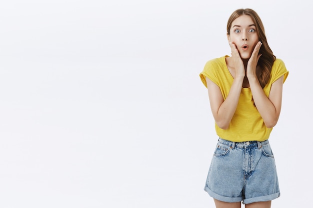 Mulher adulta encantadora e interessada espantada com camiseta amarela e shorts dobrando os lábios segurando as palmas das mãos nas bochechas de curiosidade e entusiasmo