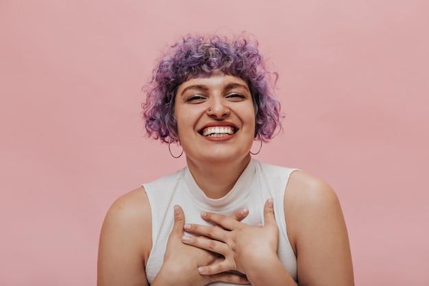 Mulher adulta sincera com cabelo roxo ondulado continua rindo. mulher com piercing posando de top branco em rosa.