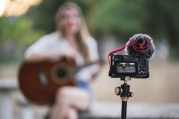 Mulher adulta tocando violão em um parque e gravando-se com uma câmera DSLR