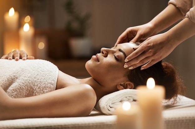 Mulher africana atraente, desfrutando de massagem de rosto no salão spa.