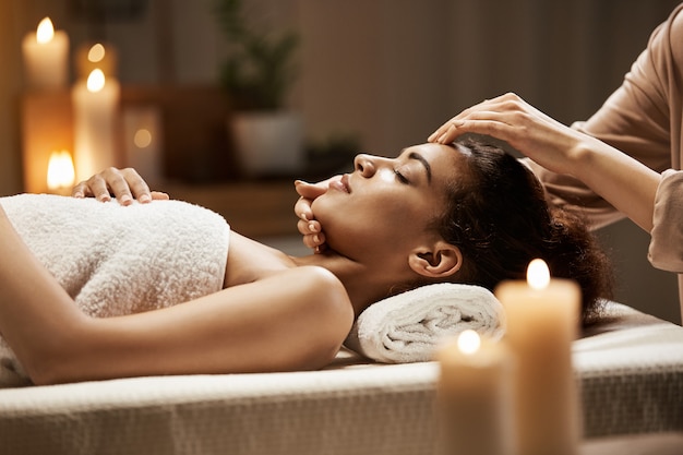 Mulher africana atraente, desfrutando de massagem de rosto no salão spa.