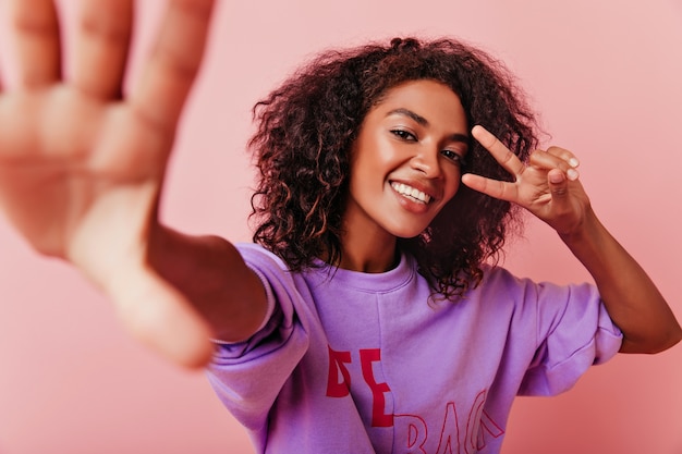 Mulher africana atraente fazendo selfie com o símbolo da paz. Retrato interior da garota da risada emocional posando em rosa.