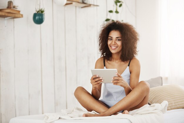 Mulher africana atrativa na roupa de noite que senta-se na cama que sorri guardando a tabuleta em casa.