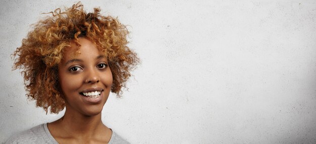 Mulher africana elegante com cabelo encaracolado e piercing no nariz, tendo uma expressão feliz e alegre, regozijando-se com algumas notícias agradáveis enquanto posava dentro de casa contra uma parede em branco com espaço de cópia para sua informação