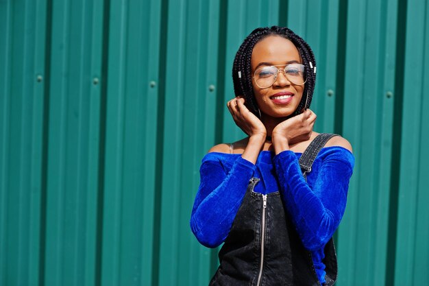 Mulher africana em óculos de saia de macacão jeans posou contra a parede de aço verde