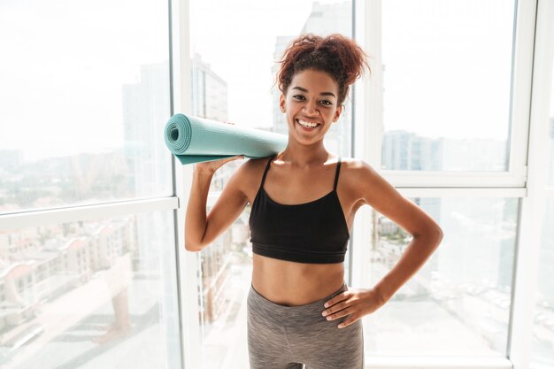 Mulher africana feliz fitness sorrindo e segurando o tapete esportivo em casa